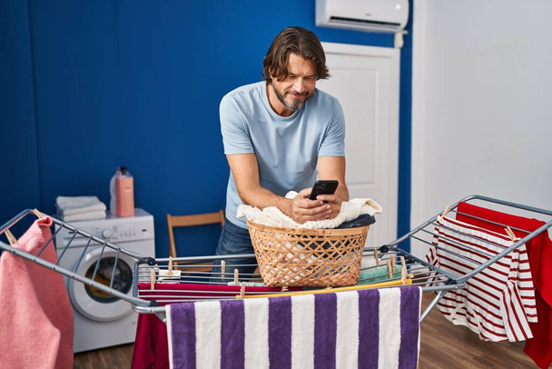 Mężczyzna w średnim wieku rozmawia na smartfonie wieszając ubrania na sznurku w pralni - Zdjęcie, obraz