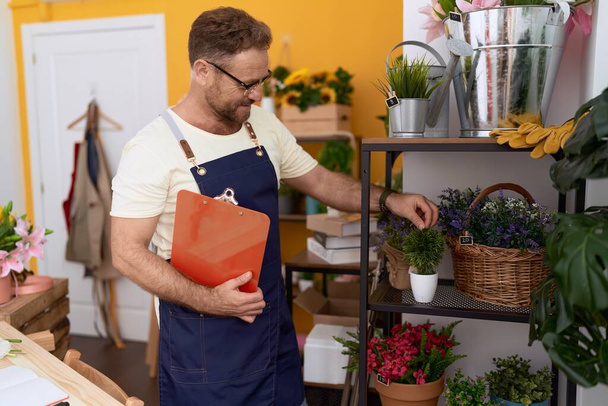 Middle age man florist touching plant holding clipboard at flower shop - 写真・画像