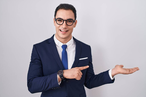 Νεαρός Ισπανός που φοράει κοστούμι και γραβάτα κατάπληκτος και χαμογελαστός στην κάμερα ενώ παρουσιάζει με το χέρι και δείχνει με το δάχτυλο.  - Φωτογραφία, εικόνα