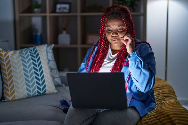 Saçları örgülü, bilgisayar laptopu kullanan, dudakları parmaklarıyla kapanan Afro-Amerikalı bir kadın. gizli ve sessiz, tabu konuşması  - Fotoğraf, Görsel