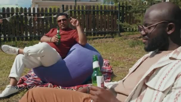 Moderní černošští a středovýchodní muži sedí uvolněně na fazolových židlích na dvorku a popíjejí pivo a mluví v letní den - Záběry, video