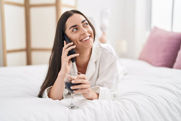 Genç İspanyol kadın akıllı telefonda konuşuyor. Yatak odasında şarap içiyor. - Fotoğraf, Görsel