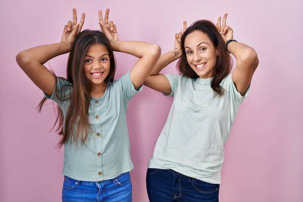 Junge Mutter und Tochter stehen auf rosa Hintergrund posieren lustig und verrückt mit den Fingern auf dem Kopf als Hasenohren, lächeln fröhlich  - Foto, Bild