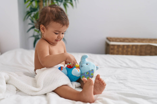 Adorable hispanic baby sitting on bed playing with elephant doll at bedroom - Photo, Image