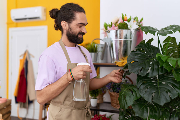 Young hispanic man florist using diffuser watering plant at flower shop - Foto, afbeelding