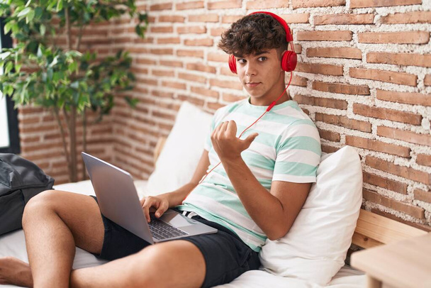 Hispanic teenager using laptop at home sitting on the bedroom pointing thumb up to the side smiling happy with open mouth  - Zdjęcie, obraz