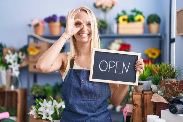 Young caucasian woman working at florist with open sign smiling happy doing ok sign with hand on eye looking through fingers  - Foto, Imagem