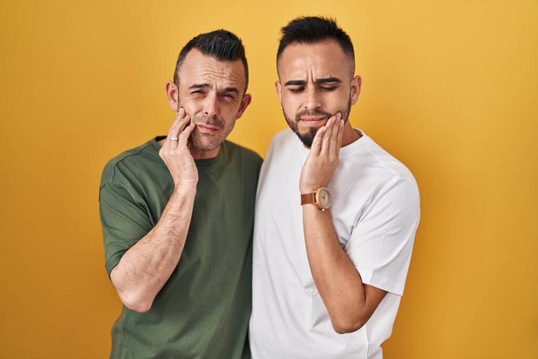 Homosexuelle Paare, die vor gelbem Hintergrund stehen, berühren den Mund mit schmerzhaftem Gesichtsausdruck wegen Zahnschmerzen oder Zahnkrankheiten an den Zähnen. Zahnarztkonzept.  - Foto, Bild