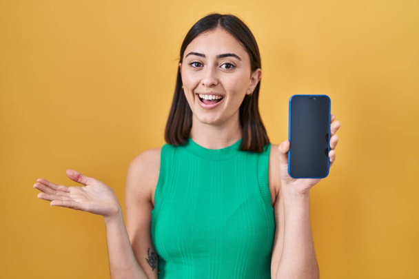 Ισπανόφωνη κοπέλα κρατώντας smartphone δείχνει οθόνη γιορτάζει επίτευγμα με χαρούμενο χαμόγελο και έκφραση νικητής με έθεσε το χέρι  - Φωτογραφία, εικόνα