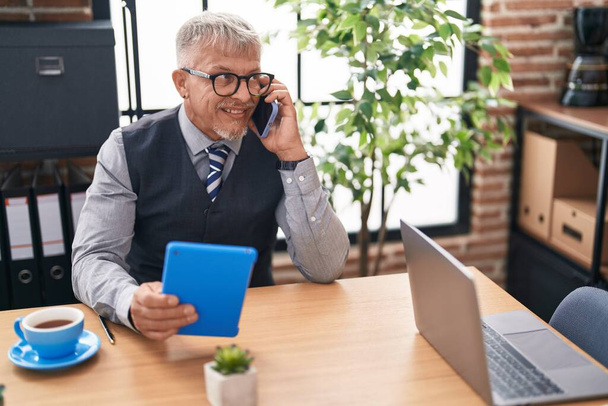Középkorú ősz hajú férfi üzleti dolgozó érintőpad beszél okostelefonon az irodában - Fotó, kép