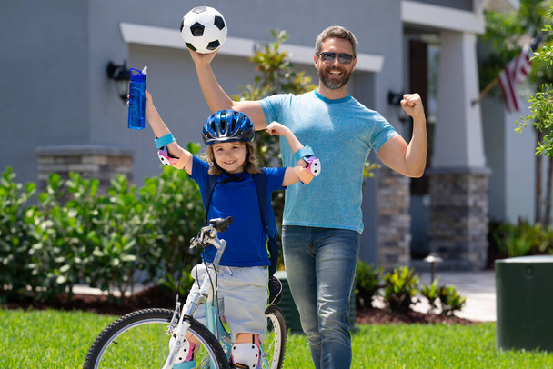 Sporty family. Excited father and son with winning gesture. Happy family. Father and son riding bike in park. Child in safety helmet with father riding bike in summer day. Father teaching son riding - Fotó, kép