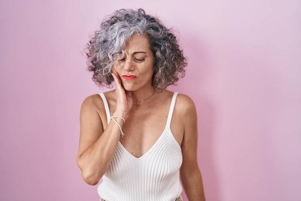 Middle age woman with grey hair standing over pink background touching mouth with hand with painful expression because of toothache or dental illness on teeth. dentist  - Foto, Imagen