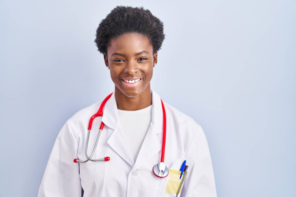 Femme afro-américaine portant l'uniforme de médecin et stéthoscope regardant positif et heureux debout et souriant avec un sourire confiant montrant des dents  - Photo, image