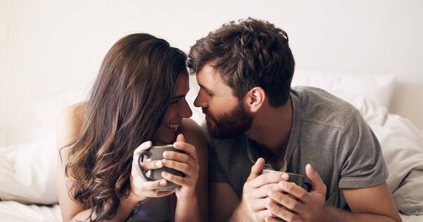 Relax, coffee and love with couple in bedroom for morning, wake up and affectionate. Happy, smile and free time with man and woman and kiss in bed at home for weekend, happiness and romance together. - Фото, изображение