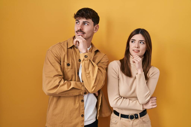 Νεαρό ζευγάρι ισπανόφωνων στέκεται πάνω από κίτρινο φόντο σκέφτεται ανησυχούν για μια ερώτηση, ανησυχούν και νευρικό με το χέρι στο πηγούνι  - Φωτογραφία, εικόνα