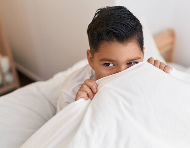 Adorável menino hispânico sentado na cama cobrindo com lençol no quarto - Foto, Imagem