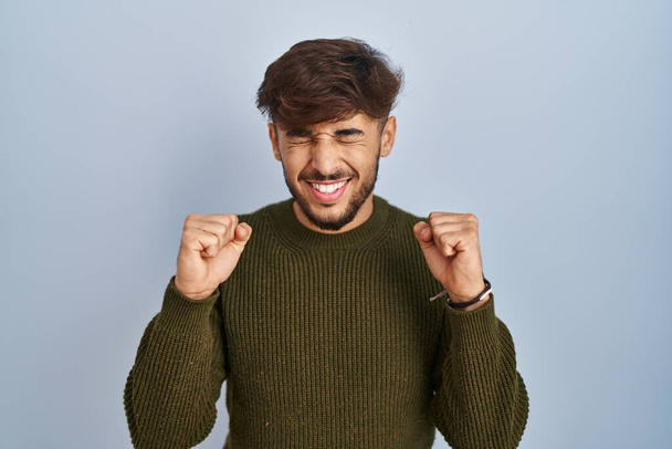 Arab man with beard standing over blue background excited for success with arms raised and eyes closed celebrating victory smiling. winner concept.  - Photo, Image