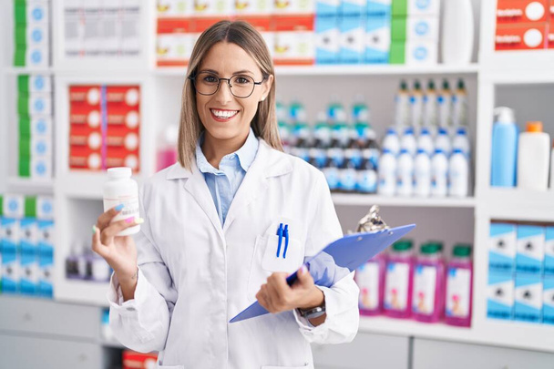 Joven hermosa mujer hispana farmacéutica sosteniendo pastillas botella lectura documento en la farmacia - Foto, imagen