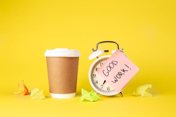 Paper cup and alarm clock with paper reminder with the inscription Good work on a yellow background isolated. - Photo, Image