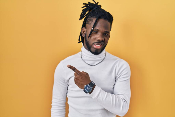 African man with dreadlocks wearing turtleneck sweater over yellow background pointing aside worried and nervous with forefinger, concerned and surprised expression  - Photo, Image