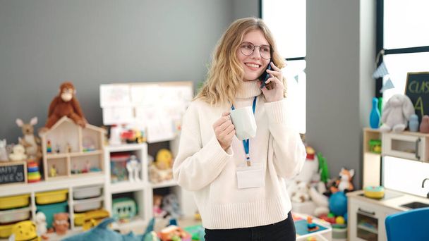 Young blonde woman preschool teacher talking on smartphone drinking coffee at kindergarten - Valokuva, kuva