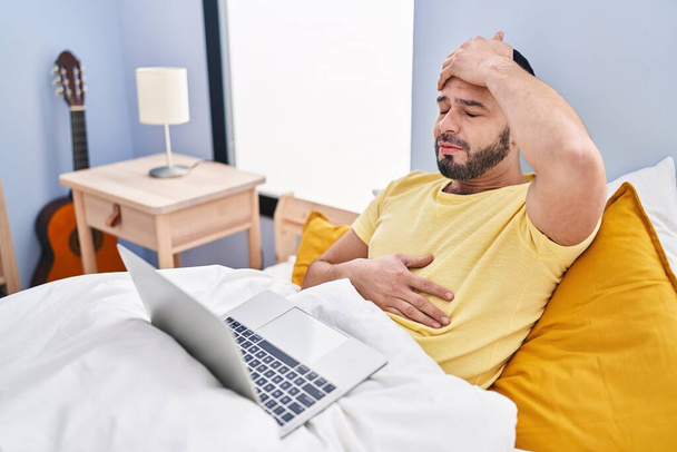 Jonge Latijnse man ziek met telegeneeskunde zittend op bed in de slaapkamer - Foto, afbeelding
