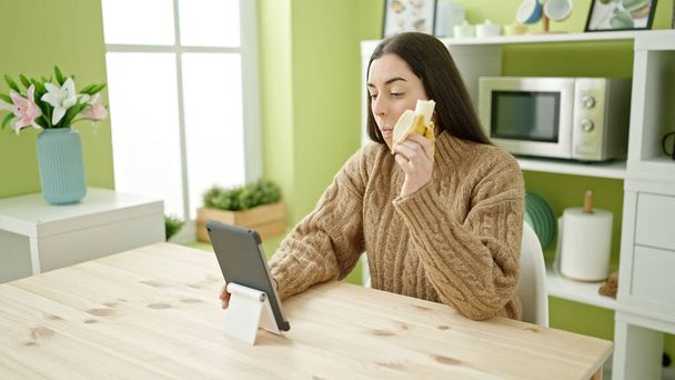 Joven hermosa mujer hispana comiendo plátano viendo video en touchpad en la cocina - Foto, imagen