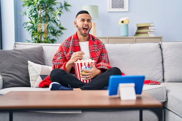 Young hispanic man eating popcorn watching movie on tablet device angry and mad screaming frustrated and furious, shouting with anger looking up.  - Foto, afbeelding