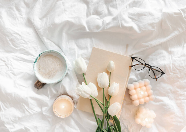 Kytice bílých tulipánů, šálek kávy, svíčky a kniha v bílé posteli, pohled shora. - Fotografie, Obrázek