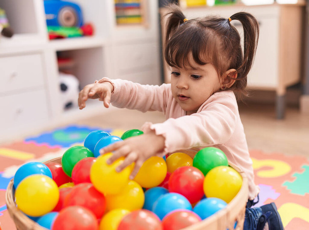 Adorable hispanic girl playing with balls sitting on floor at kindergarten - 写真・画像