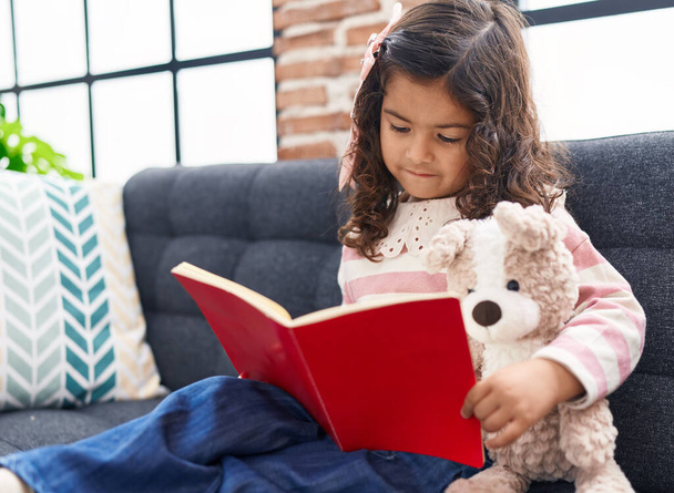 Adorable hispanic girl reading book sitting on sofa at home - Foto, immagini