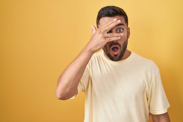 Handsome hispanic man standing over yellow background peeking in shock covering face and eyes with hand, looking through fingers with embarrassed expression.  - Photo, Image