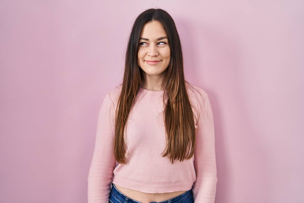 Mladá brunetka žena stojící nad růžovým pozadím s úsměvem dívá do strany a zírá pryč myšlení.  - Fotografie, Obrázek