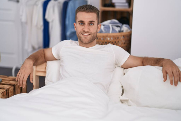 Jeune homme caucasien souriant confiant assis sur le lit dans la chambre - Photo, image