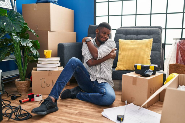 Αφροαμερικάνος που κάθεται στο πάτωμα στο νέο σπίτι αγκαλιάζοντας τον εαυτό του χαρούμενο και θετικό, χαμογελώντας με αυτοπεποίθηση. αυτοαγάπη και αυτοφροντίδα  - Φωτογραφία, εικόνα