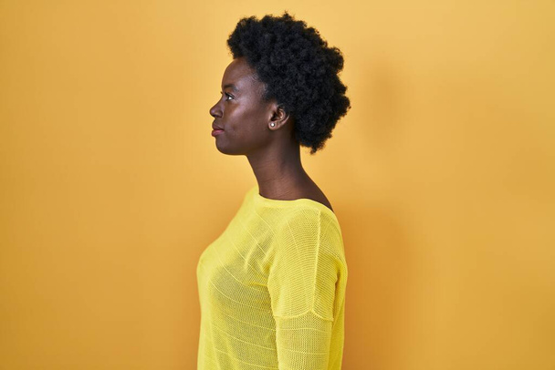 Африканская молодая женщина стоит над желтой студии глядя в сторону, расслабиться профиль позировать с естественным лицом и уверенной улыбкой.  - Фото, изображение