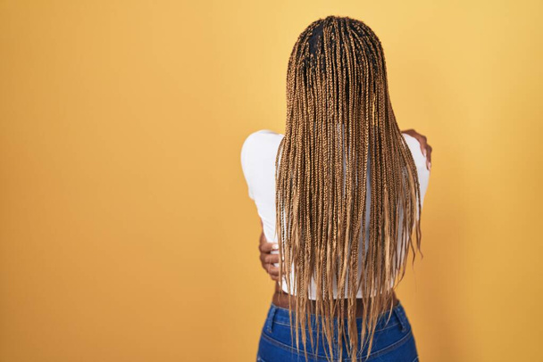 African american woman with braided hair standing over yellow background hugging oneself happy and positive from backwards. self love and self care  - Foto, Imagem