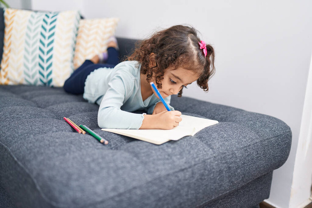 Adorable chica hispana dibujando en un cuaderno tumbada en un sofá en casa - Foto, Imagen