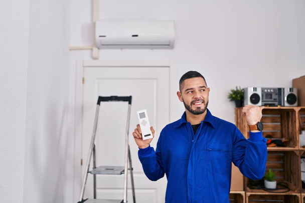 Hispanische Reparaturfirma arbeitet mit Klimaanlage und zeigt mit dem Daumen zur Seite, lächelt glücklich mit offenem Mund  - Foto, Bild