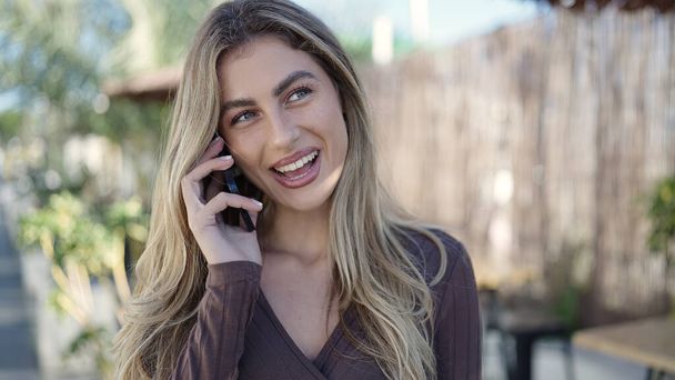 Молодая блондинка улыбается уверенно разговаривая на смартфоне на террасе кафе - Фото, изображение