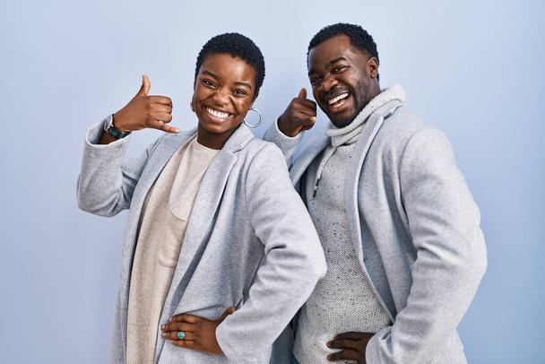 Nuori afrikkalainen amerikkalainen pariskunta seisoo sinisen taustan päällä hymyillen ja tehden puhelimen elettä käsin ja sormin kuin puhuen puhelimessa. käsitteiden välittäminen.  - Valokuva, kuva