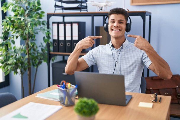 Mladý hispánec pracující v kanceláři ve sluchátkách, vesele se usmívající a ukazující prsty a ústy. koncepce stomatologického zdraví.  - Fotografie, Obrázek