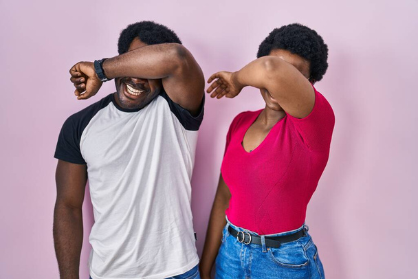 Genç Afro-Amerikan çifti pembe arka planda dikiliyor. Gözleri kollarıyla kaplıyor. Neşeli ve komik gülümsüyor. kör kavram.  - Fotoğraf, Görsel