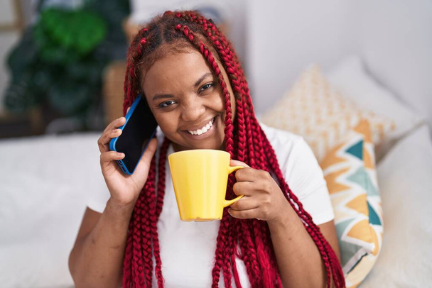 Afrika kökenli Amerikalı kadın akıllı telefondan konuşuyor. Yatak odasında kahve içiyor. - Fotoğraf, Görsel