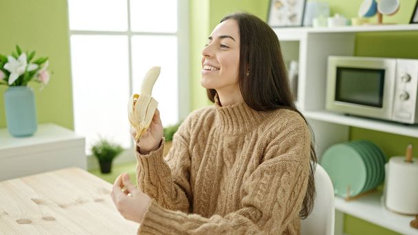 Giovane bella donna ispanica mangiare banana seduto sul tavolo in cucina - Foto, immagini