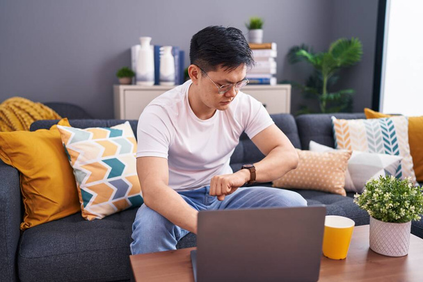 Jeune homme asiatique à l'aide d'un ordinateur portable à la maison assis sur le canapé vérifier l'heure sur la montre-bracelet, détendu et confiant  - Photo, image