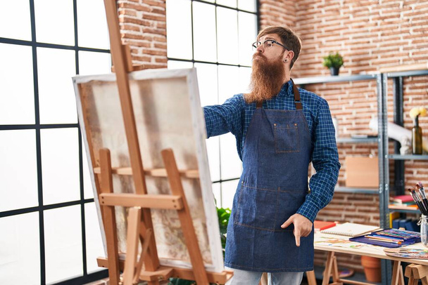 Jovem ruiva homem artista olhando desenhar com expressão de dúvida no estúdio de arte - Foto, Imagem