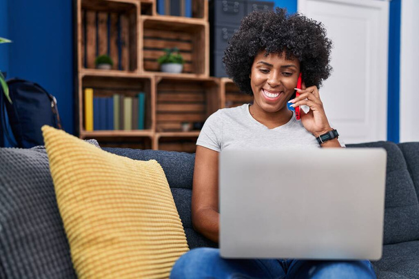 African american woman talking on smartphone using laptop at home - Фото, зображення