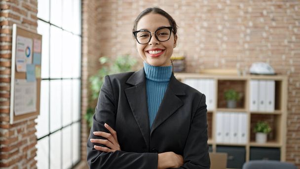 Νεαρή όμορφη Ισπανίδα εργαζόμενη επιχειρηματίας χαμογελώντας με σταυρωμένα χέρια στο γραφείο - Φωτογραφία, εικόνα