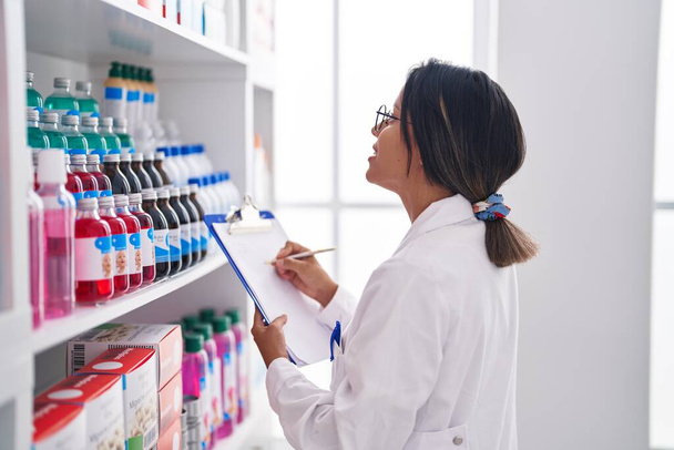 Joven farmacéutica hispana escribiendo en documento mirando estanterías en farmacia - Foto, Imagen